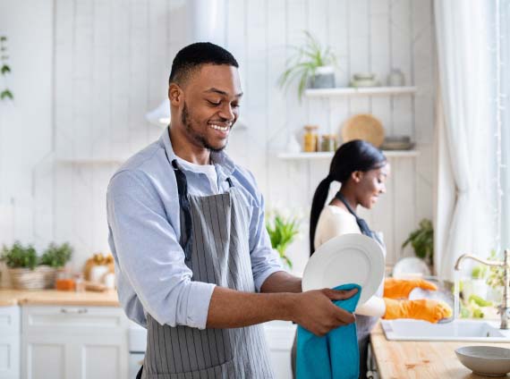 man drying plate with a smile on his face while woman washes dishes in the background